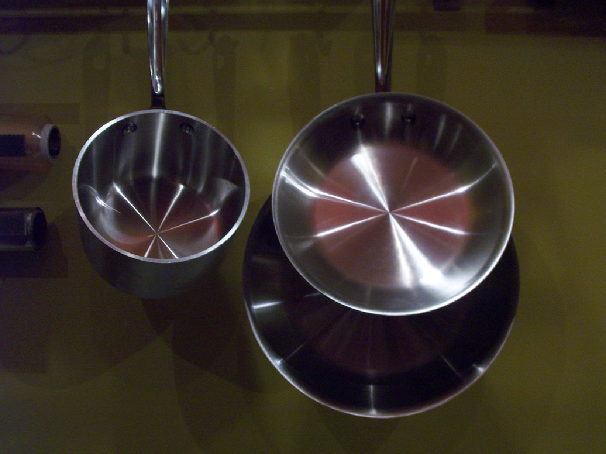All-Clad stainless steel fry pan and saucepan hanging from a pot rack, polished bonded-steel interiors catching the light