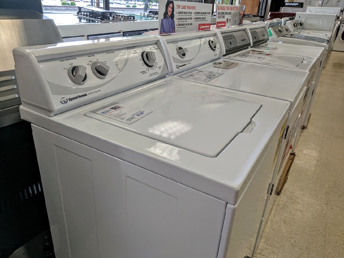 Row of white Speed Queen Commercial Heavy Duty top-load washers with mechanical dials in an appliance showroom